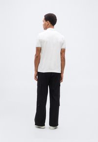 Λευκό πολο με τόνους τιρκουάζ, συνδυασμένο με φαρδιά μαύρη παντελόνια cargo με τσέπες. Το μοντέλο φαίνεται από πίσω.