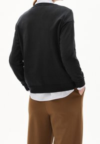 Sweatshirt gris foncé avec un col rond, des poignets et un ourlet côtelés, associé à un pantalon marron, vu de dos avec une chemise blanche en dessous.