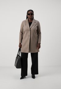 Manteau en laine texturé marron clair avec un col à revers, deux boutons et poches avant, associé à un haut noir et un pantalon large. Sac fourre-tout noir.