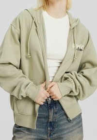 Felpa verde chiaro con zip, corde di regolazione, polsini a coste e tasca a marsupio, indossata sopra una t-shirt bianca e jeans blu.
