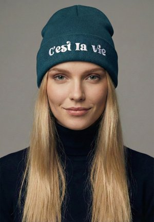 Cappello a cuffia di colore turchese lavorato a maglia con la scritta "C'est la Vie" ricamata in bianco. Presenta una texture a coste e una vestibilità casual, indossato con un dolcevita nero.