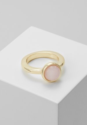 Bague en or avec une pierre ronde, lisse et rose pâle sertie dans un chaton, exposée sur un socle blanc sur fond gris.
