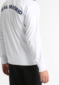 Witte jas met "REAL MADRID" in marineblauw gedrukt op de bovenrug. Ribgebreide manchetten en zoom, zachte textuur, casual pasvorm.