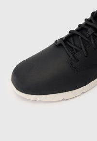Zapato de cuero negro con textura lisa, punta redondeada y suela de goma blanca en contraste. Presenta cordones negros y un diseño minimalista.