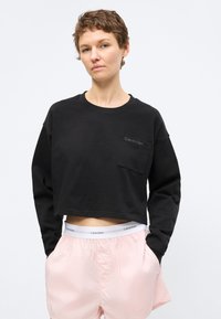 Vrouw met kort haar draagt een zwart cropped shirt met lange mouwen en lichtroze korte broek met zichtbare Calvin Klein tailleband, handen in de zakken.