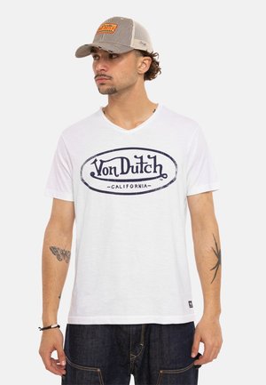 Homme portant un t-shirt blanc Von Dutch California, une casquette en mesh Von Dutch, un jean foncé, avec des tatouages de papillon et d’araignée sur les bras.