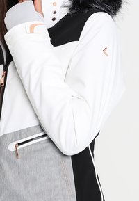 Veste d'hiver blanche, grise et noire avec fermeture éclair, boutons et logo en or rose, dotée d'une capuche bordée de fourrure noire.