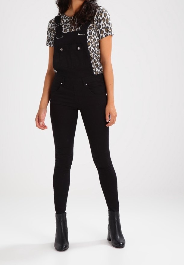 Femme portant une salopette noire ajustée, un t-shirt à manches courtes imprimé léopard, et des bottines noires, debout sur fond blanc.