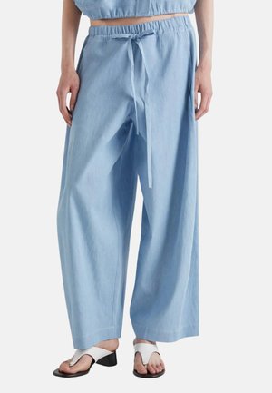 Vrouw draagt losse lichtblauwe broek met trekkoord en witte sandalen met hak, tilt haar tenen op, staand tegen een effen achtergrond.