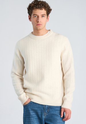Junger Mann mit lockigem Haar trägt einen cremefarbenen, grob gestrickten Pullover und blaue Jeans, steht mit einer Hand in der Tasche vor hellem Hintergrund.