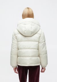 Dunjakke i lys kremfarge, med hette, quiltet design og elastiske mansjetter, kombinert med mørkerøde bukser.