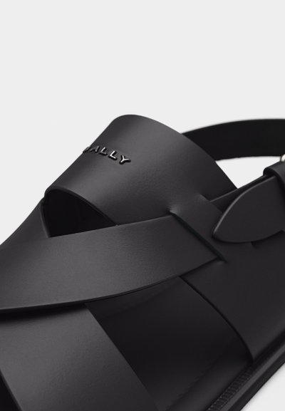 Μαύρο δερμάτινο σανδάλι με φαρδιές διασταυρούμενες λουριά και ανάγλυφο όνομα μάρκας στο επάνω λουρί, με ρυθμιζόμενο πίσω τοκά.