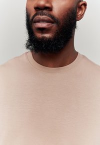 Beige short-sleeve t-shirt with a crew neckline, smooth texture, and minimal stitching details, shown on a person with a black beard.