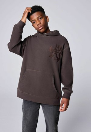 Teenager trägt dunkelbraunen Hoodie mit Fronttasche und schwarze Jeans, kratzt sich am Kopf und steht vor einem einfarbigen hellgrauen Hintergrund.