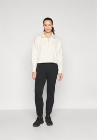 Hvid zip-up sweatshirt med høj krave, parret med sorte tapered sweatpants og sorte sneakers, stående på en ensfarvet baggrund.