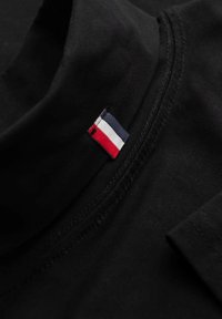 Schwarzes Polohemd mit kurzen Ärmeln, ausgestattet mit einem kleinen rotem, weißen und blauen gestreiften Logoetikett am Kragen. Glatte Stoffoberfläche.