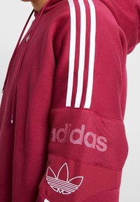 Maroon hoodie med vit tre-strängad design på ärmarna, meshnätspanel på armen och grafisk logotypdetalj på ärmen. Justerbar dragsko i huvan.