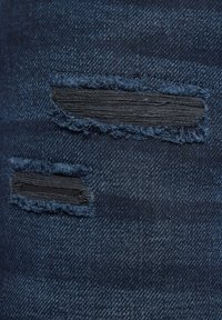 Granatowe dżinsy z denimu z dwoma przetartymi łatami z czarnym materiałem pod spodem, z frędzlami na brzegach i teksturowaną powierzchnią.