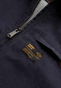 Marineblå stofjakke med guld lynlås, stribet indre for og en sort label med gul tekst, der angiver brand og plejeinformation.