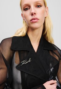 Sort mesh trenchcoat med bred krave, sølv "K" broche accent, med en struktureret, lagdelt design og tekstureret stof.