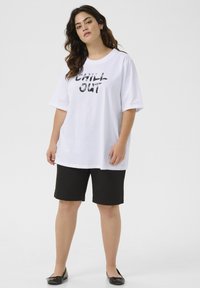 Camiseta blanca de algodón con el texto "CHILL OUT" en una fuente negra en negrita, combinada con pantalones cortos negros. El modelo lleva zapatos planos negros. Diseño simple y relajado.