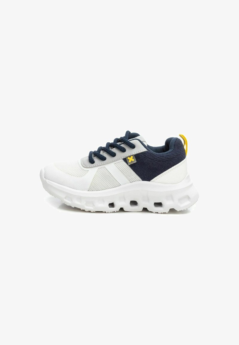 Zapatilla deportiva blanca con acentos de tela azul marino, parte superior de malla, suela de goma texturizada y bucle en el talón amarillo. Presenta cordones e inserciones elásticas.