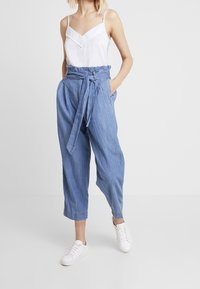 Světle modré chaotické kalhoty s vysokým pasem a páskem na zavázání, široké nohavice a kapsy po stranách, spárované s bílou pruhovanou tílkem.