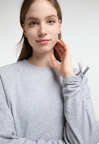 Világosszürke pulóver bordázott nyakkal és ráncolt mandzsettával. Puha textúra és diszkrét minta. A modell az arcához emelt kézzel pózol.