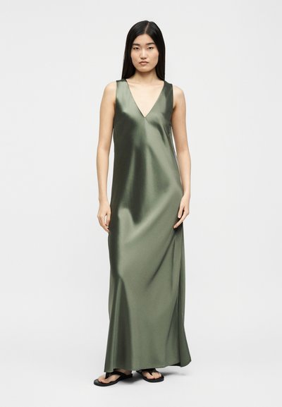 Γυναίκα που στέκεται φορώντας ένα αμάνικο μάξι φόρεμα από σατέν σε ελαιί πράσινο με V-λαιμόκοψη και μαύρα σανδάλια σε λευκό απλό φόντο.