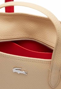 Sac en cuir beige avec une surface texturée, doté d'un intérieur rouge et d'une fermeture zippée. Logo Lacoste à l'avant.
