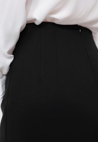 Zwarte op maat gemaakte rok met twee voorplooien, gemaakt van een gladde stof, gecombineerd met een vloeiende witte blouse die gedeeltelijk zichtbaar is.