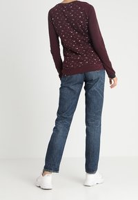 Burgundy långärmad topp med ett mönstrat bakstycke, i kombination med blå raka jeans och vita sneakers. Texturen ser mjuk och avslappnad ut.