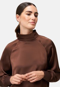 Brązowy sweter z wysokim kołnierzem wykonany z gładkiego materiału, z rękawami raglanowymi i marszczonymi mankietami, o minimalistycznym designie i subtelnym połysku.