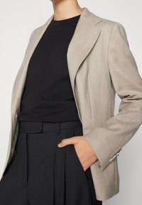 Beige bleiseri, jossa on taskut ja kaulukset, päällä musta T-paita. Kankaan pinta on teksturoitu, ja siinä on muotoillut hihat sekä kapea istuvuus.
