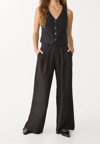 Czarna kamizelka z dekoltem w serek, pięcioma guzikami i dwoma bocznymi kieszeniami, w połączeniu z luźnymi czarnymi spodniami i spiczastymi butami.