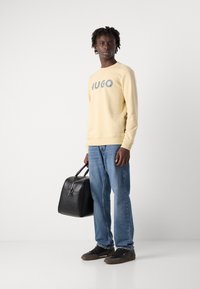 Gul genser med "HUGO" trykket i grått, kombinert med blå jeans. En svart skinnveske og svarte sko fullfører antrekket.