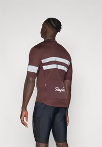 Krátký rukáv cyklistického dresu v burgundské barvě s reflexními pruhy a kapsou na zádech. Černé šortky s logem, hladká textura po celém povrchu.