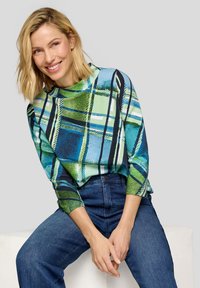 Lächelnde Frau mit blondem Haar, die ein langärmliges kariertes Oberteil in Grün und Blau sowie blaue Jeans trägt und auf einem weißen Block vor grauem Hintergrund sitzt.