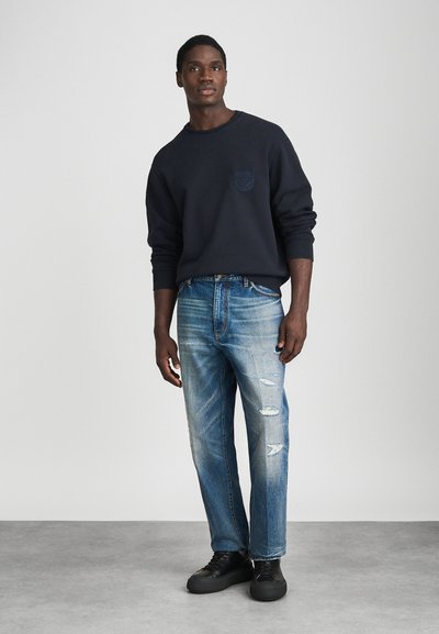 Άνδρας στέκεται με μαύρη φούτερ μπλούζα με λογότυπο στο στήθος, ξεθωριασμένα σκισμένα μπλε τζιν και μαύρες δερμάτινες αθλητικές παπουτσιάρες σε γκρι πάτωμα.