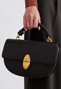 Main tenant un sac à main en cuir noir avec une poignée tressée et un fermoir ovale en or, sur un fond en tissu sombre.