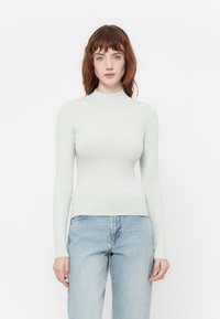 Lys grønn ribbestrikket langermet topp med høy hals, tettsittende design og subtil logo detalj på brystet, sammen med blå denimbukser.