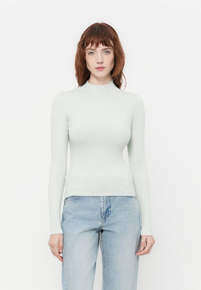 Lys grønn ribbestrikket langermet topp med høy hals, tettsittende design og subtil logo detalj på brystet, sammen med blå denimbukser.