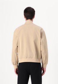 Beige bomber jacket with a smooth texture, ribbed cuffs and hem, and a relaxed fit. Black pants with side accents complete the outfit.