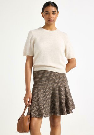 Cremefarvet kortærmet strikket sweater parret med en brunternet flare-nederdel. Modellen holder en lille brun håndtaske. Teksturen virker blød og let.