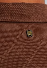 Bruine textuurstof met een geruit patroon en een klein zwart label met gouden tekst "PME LEGEND" dat met een bruine draad is bevestigd.
