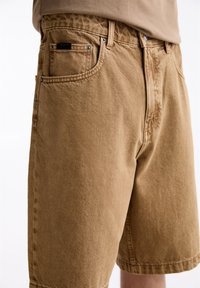 Pantalones cortos de algodón marrón con un corte recto, que cuentan con bolsillos traseros, detalles de costura sutiles y cierre de cremallera. Tela texturizada con un acabado suave.