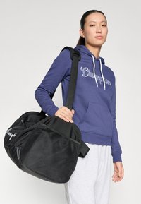 Black duffle bag with a textured surface, a mesh side panel, and a thick black strap. Model wears a purple hoodie with a kangaroo pocket.