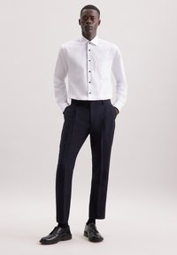 Witte lange mouwen overhemd met knoopsluiting aan de voorkant en een enkele borstzak, gecombineerd met op maat gemaakte donkere broek en zwarte veterschoenen.