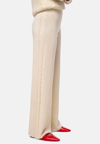 Pantalon large crème avec des détails en tricot à cable sur les côtés, associé à des chaussures à bout pointu rouges, fabriquées en tissu doux.