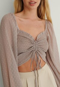 Grå, strukturerad crop top med en smockad design, som har långa, genomskinliga ärmar och en knuten detalj fram. Mjuk, lättviktig tyg med subtila mönster.